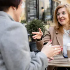 Dwie kobiety rozmawiają przy stoliku. Jedna z nich, z blond włosami, z uśmiechem gestykuluje, opowiadając o tym, jak rozmawiać z osobą w manii.
