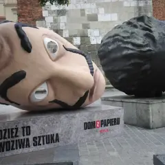Dwie gigantyczne głowy, jedna jasna, druga ciemna, sugerują, z jakiego materiału można wykonać rzeźbę. Napis "Dom z papieru" informuje o pochodzeniu.