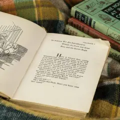 Otwarta książka z ilustracją Kubusia Puchatka i Christophera Robina na schodach. W tle stos książek A.A. Milne'a.