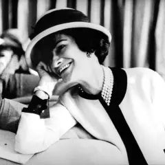 Uśmiechnięta kobieta w kapeluszu i perłach, z dłonią opartą o policzek. Ikona stylu, Coco Chanel, w swoim charakterystycznym, eleganckim stroju.