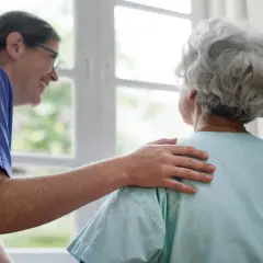 Opiekunka z czułością kładzie dłoń na ramieniu starszej pani. W jej spojrzeniu widać troskę, jakby babcia wchodziła w rolę matki.