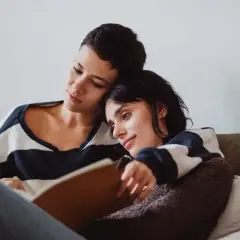 Dwie kobiety przytulone na kanapie, jedna czyta, druga opiera głowę na jej ramieniu. W tle widać książki o lesbijkach.