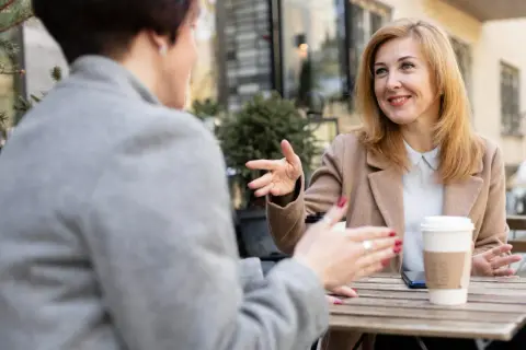 Dwie kobiety rozmawiają przy stoliku. Jedna z nich, z blond włosami, z uśmiechem gestykuluje, opowiadając o tym, jak rozmawiać z osobą w manii.