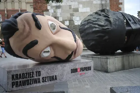 Dwie gigantyczne głowy, jedna jasna, druga ciemna, sugerują, z jakiego materiału można wykonać rzeźbę. Napis "Dom z papieru" informuje o pochodzeniu.