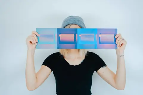 Osoba w czapce i czarnej koszulce trzyma przed twarzą panel z czterema prostokątami w odcieniach niebieskiego i różowego. Inspiracja do studiów artystycznych kierunki.