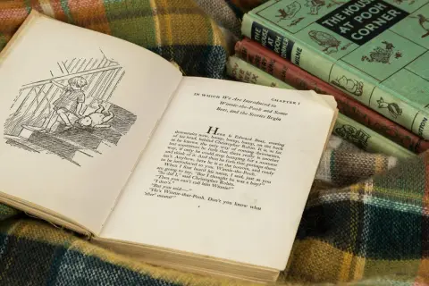 Otwarta książka z ilustracją Kubusia Puchatka i Christophera Robina na schodach. W tle stos książek A.A. Milne'a.