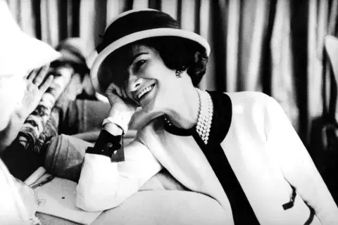 Uśmiechnięta kobieta w kapeluszu i perłach, z dłonią opartą o policzek. Ikona stylu, Coco Chanel, w swoim charakterystycznym, eleganckim stroju.