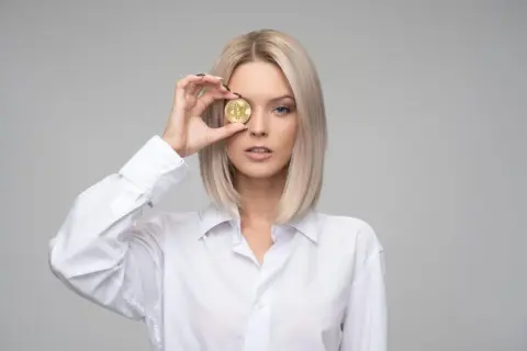 Blondynka w białej koszuli trzyma monetę bitcoin przed okiem, czując zazdrość, że ktoś ma lepiej.