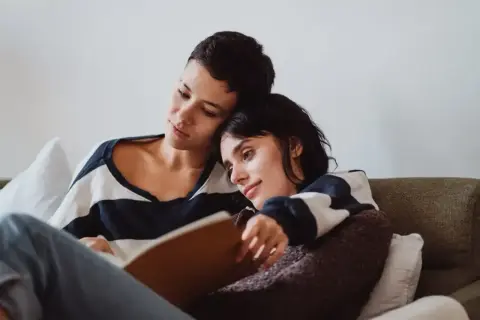 Dwie kobiety przytulone na kanapie, jedna czyta, druga opiera głowę na jej ramieniu. W tle widać książki o lesbijkach.